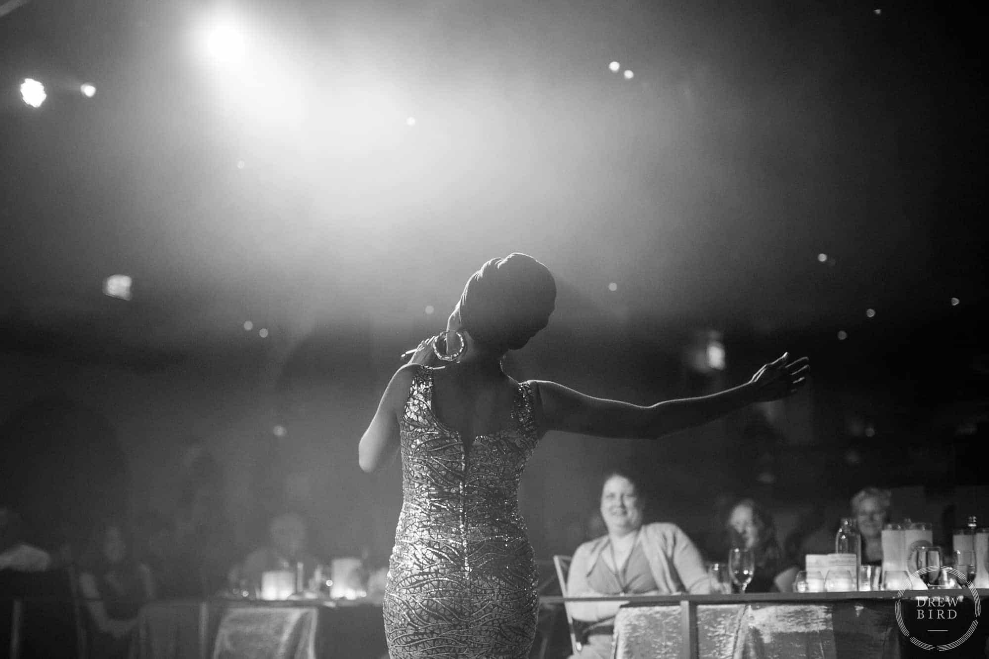 Woman with back turned to the camera singing to an audience seated at tables. Part of a corporate photography event, photographed by Drew Bird in San Francisco.