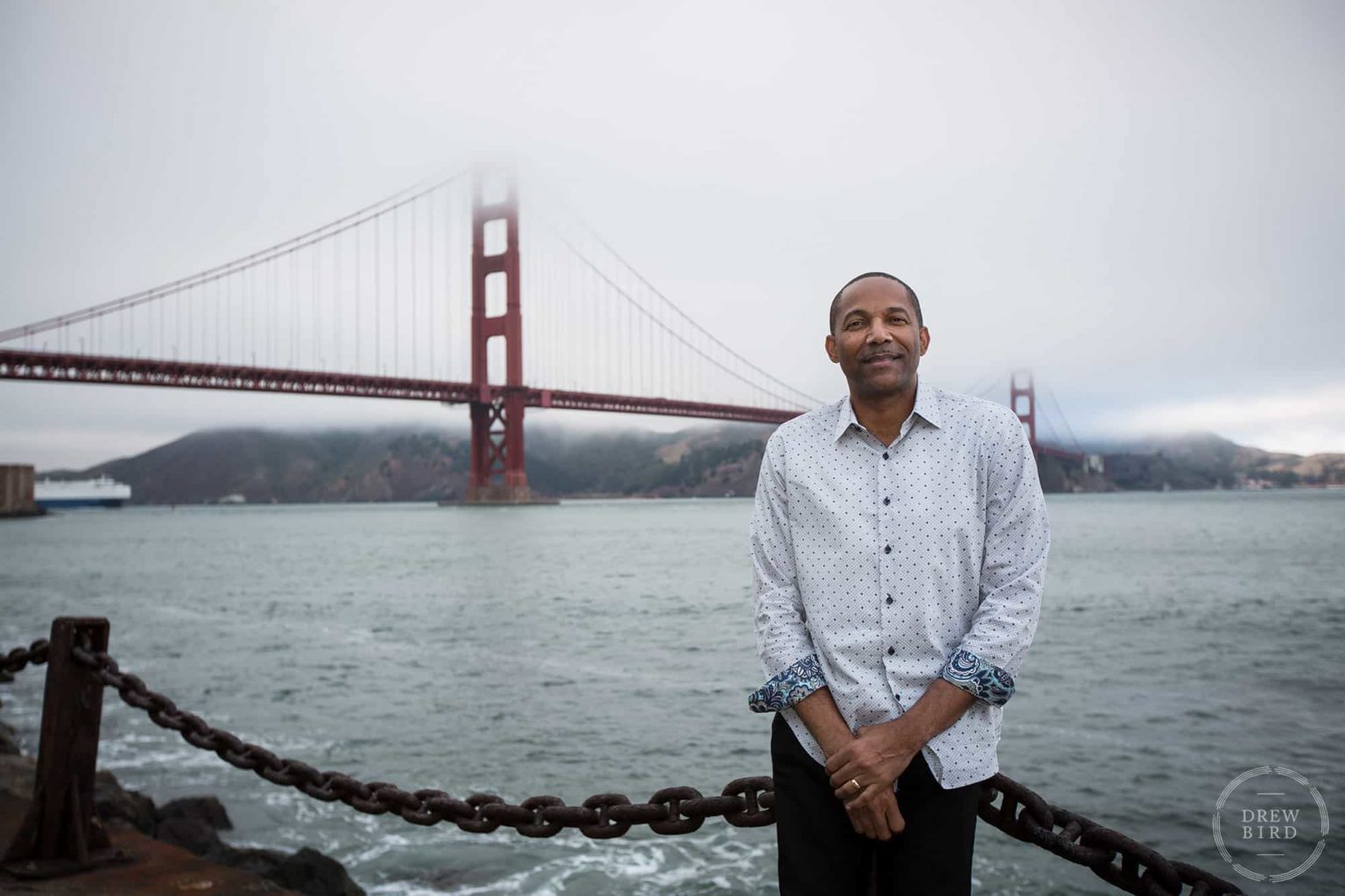 A man leans on a chain fence with Golden Gate Bridge. San Francisco editorial portrait photographer Drew Bird.