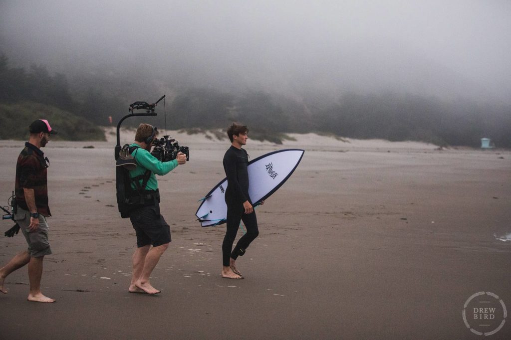 Film crew filming a man with a surfboard on beach in Stinson Beach. San Francisco movie set unit still photographer Drew Bird